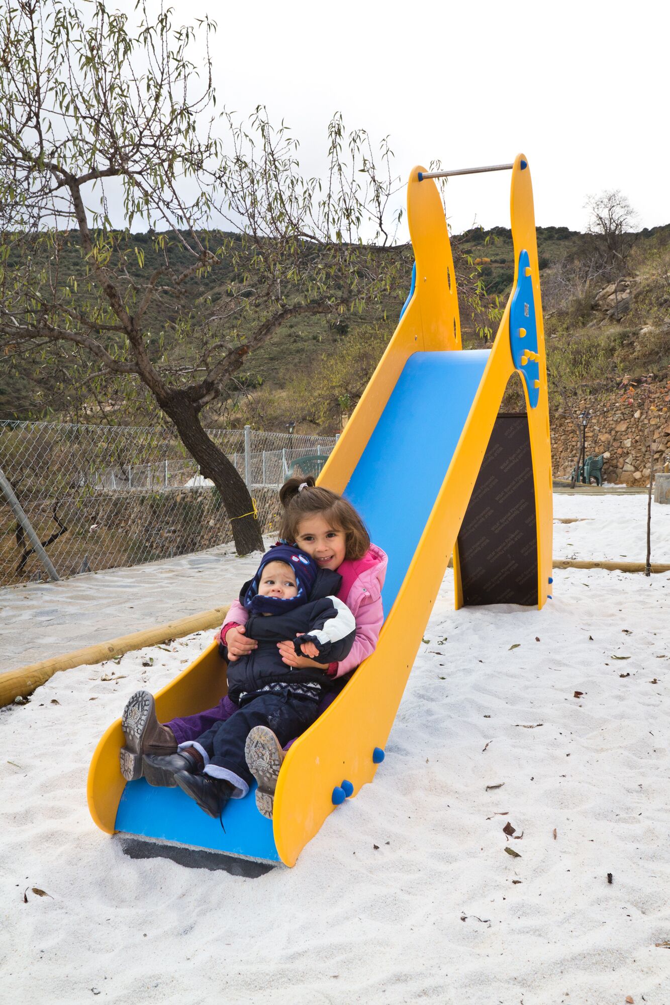 Two children on a yellow and blue slide in a snowy outdoor area.