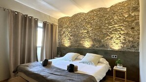 A bedroom with a stone wall, a bed with a grey blanket, a wooden nightstand, and a window with curtains. - Rural House La Casa dels Avis (Ullà)