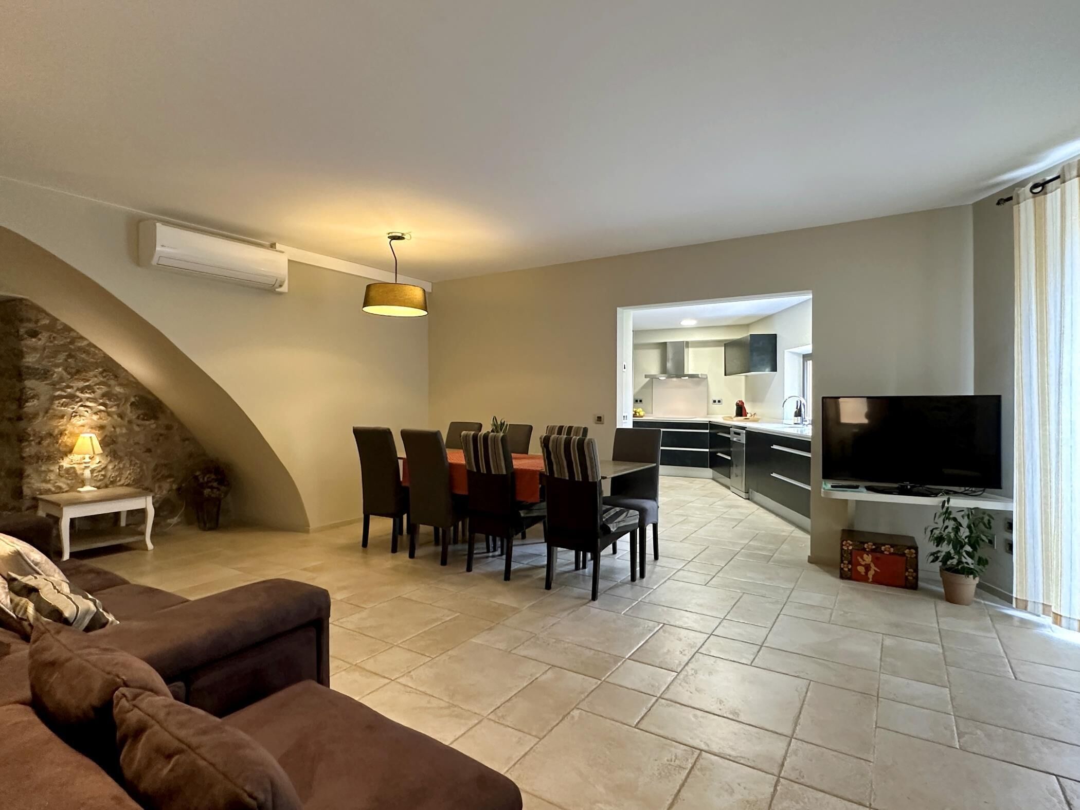 A modern living room with a sofa, dining table, chairs, and a TV. There is a kitchen visible in the background.