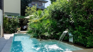 Une piscine agrémentée d’une cascade, entourée d’une végétation luxuriante, avec un bâtiment visible au loin.