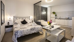 Une chambre moderne avec un grand lit, des tables de chevet, une petite table avec un vase, et une vue sur la cuisine adjacente.