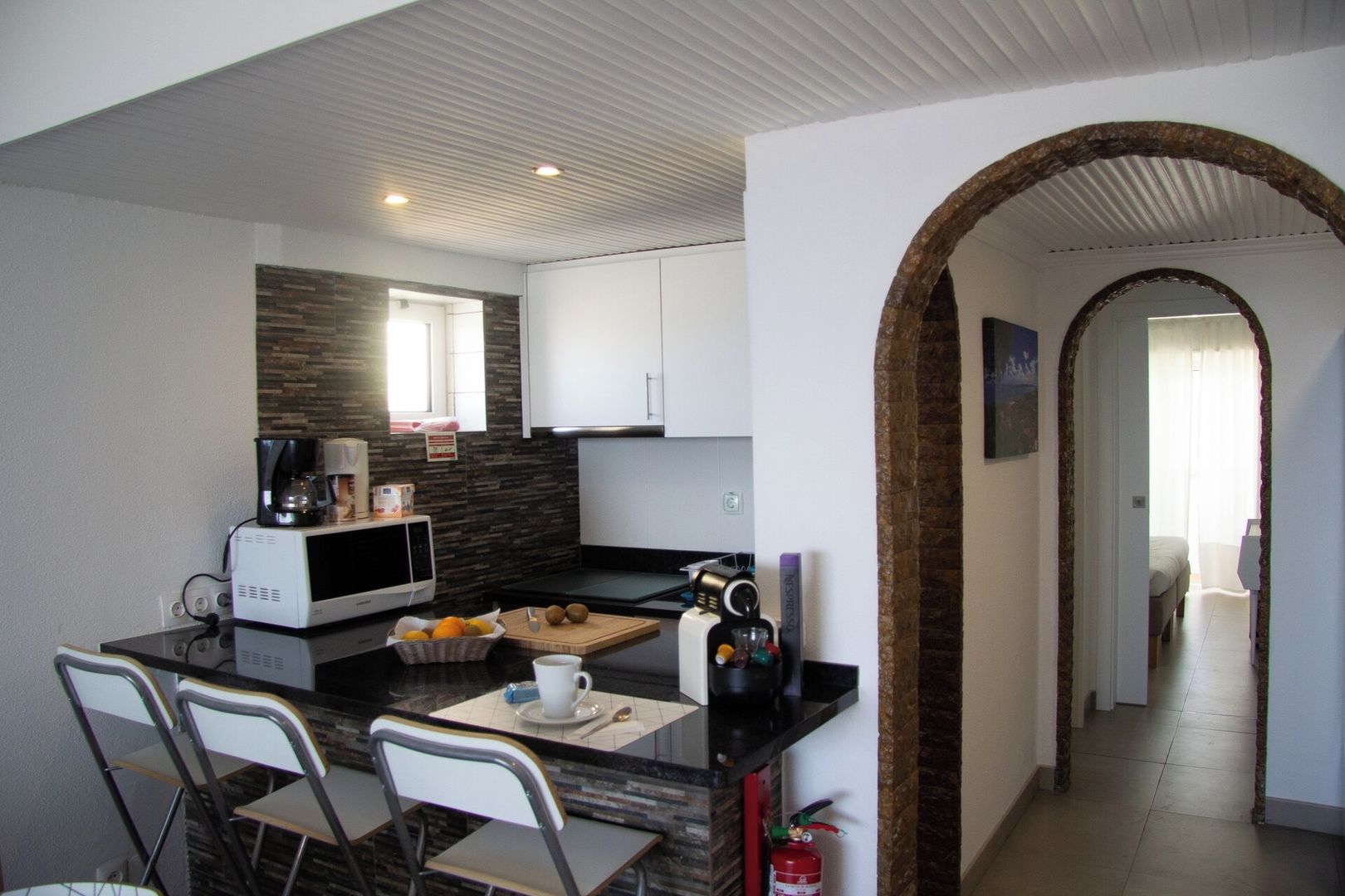 A compact kitchen with a microwave, coffee maker, and a fruit bowl on the counter. There are two chairs and a small dining table.