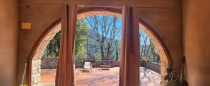 Un portail en arc mène à un patio extérieur doté de murs en pierre, de meubles en bois et offrant une vue sur les arbres et les collines.