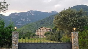 A gated entrance to a building with a stone base and metal gate, surrounded by lush greenery and mountains in the background. - Self catering Can Servosa for 11 people (Sales de Llierca)