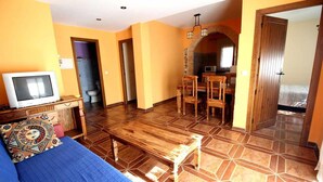 Quarto com piso de madeira, cama com cobertor azul, mesa de madeira, televisão e pequena área de cozinha com micro-ondas.