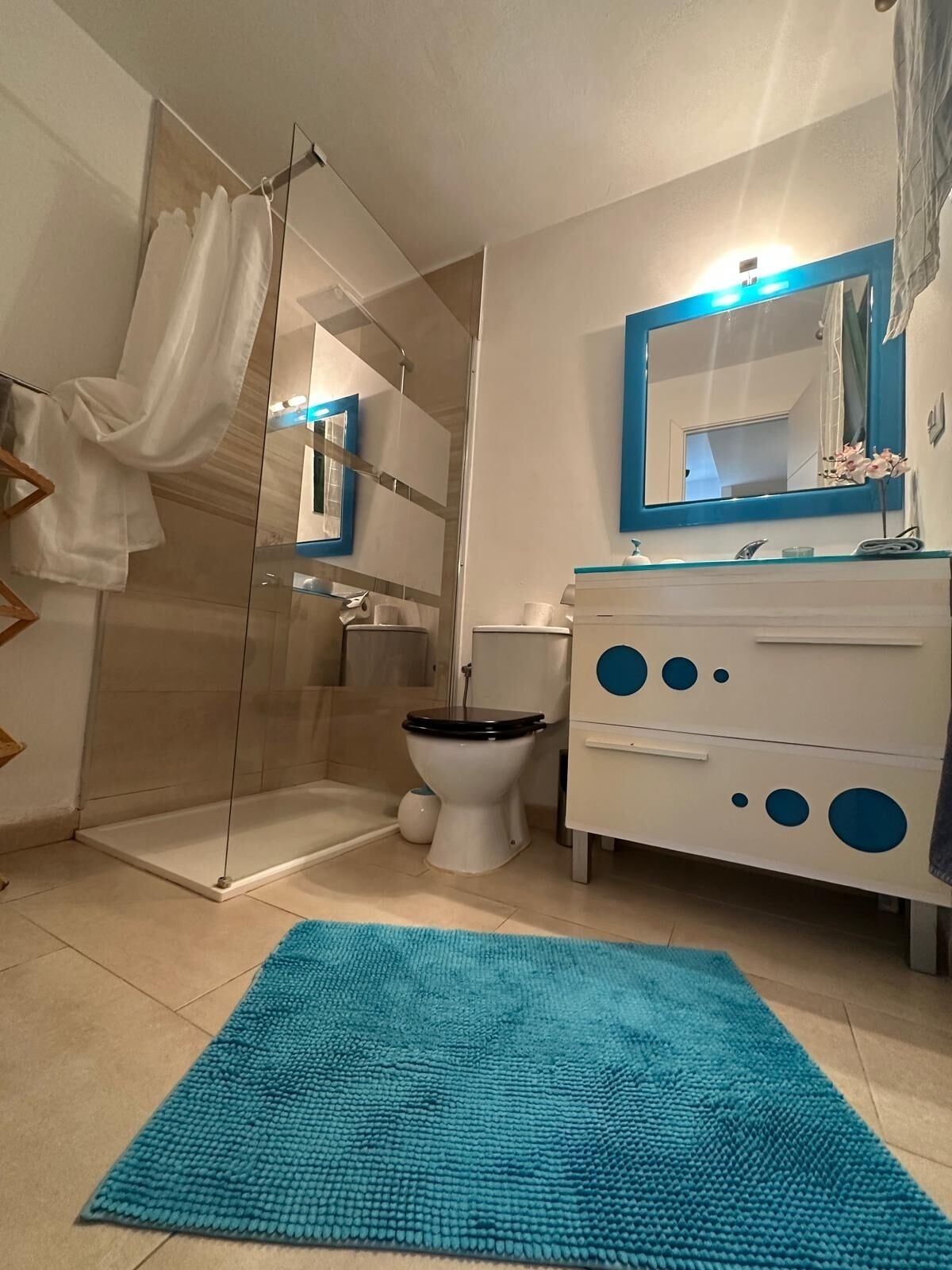 A bathroom with a glass shower enclosure, a white toilet, a blue-framed mirror, and a white vanity with circular cutouts.