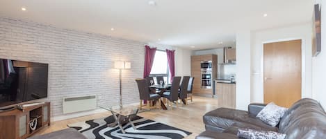 Un salon moderne avec un canapé en cuir, une table basse en verre et un téléviseur à écran plat. On y trouve également un coin repas avec une table et des chaises, et une cuisine à l’arrière-plan.