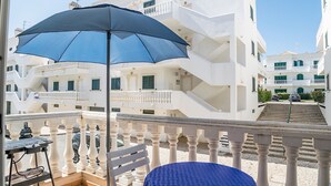 Un balcón con una mesa y una silla, una sombrilla azul y un edificio blanco con balcones y escaleras.