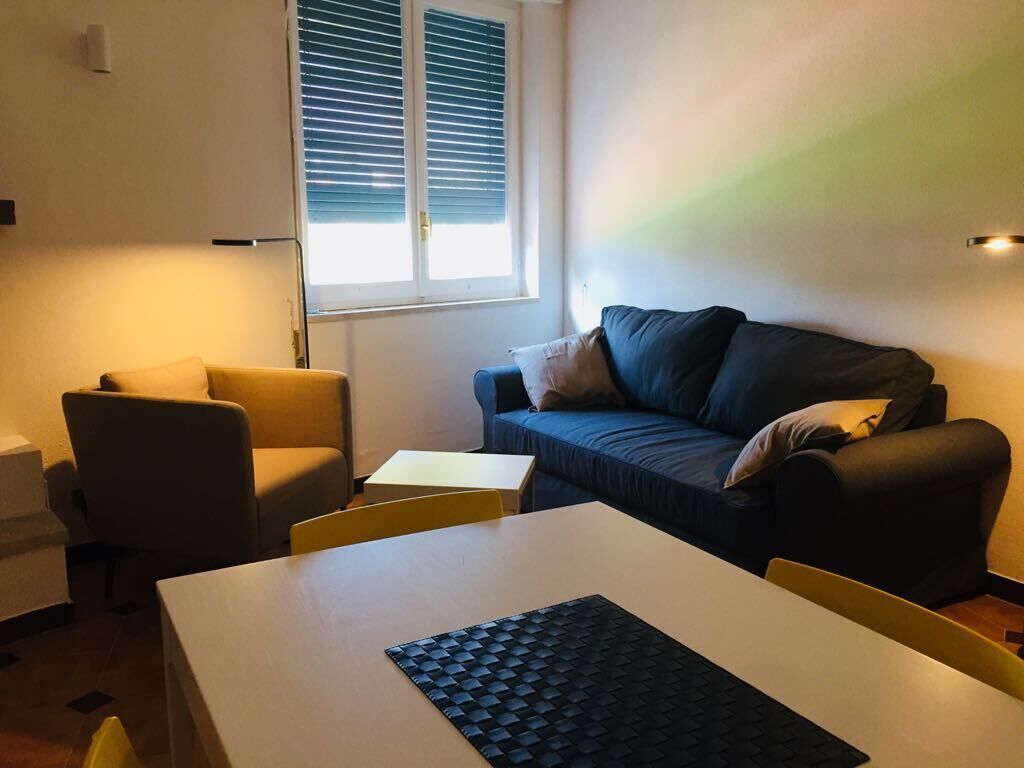 A small, well-lit room with a sofa, a table, and a chair. There is a window with blinds, a lamp on the wall, and a small table with a keyboard.