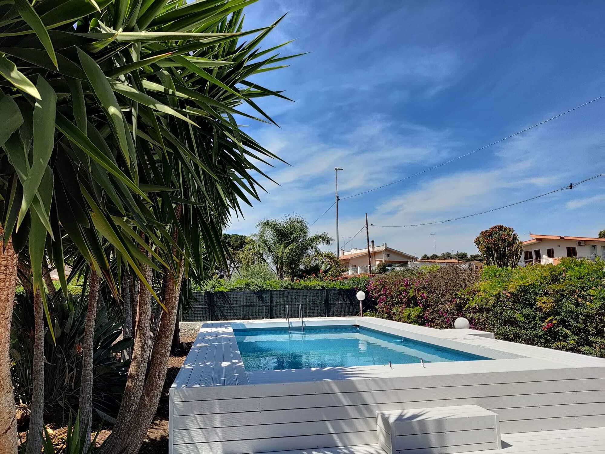 Una piscina rettangolare con bordo in legno, immersa nel verde e con un cielo azzurro limpido.