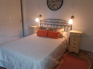 Una cama bien hecha con edredón blanco y almohadas naranjas, dos mesitas de noche con lámparas y un reloj de pared.