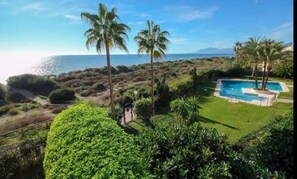 A pool surrounded by lush greenery and palm trees, with a view of the ocean and mountains in the background.