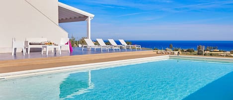 Un espace aménagé au bord de la piscine, avec des chaises longues et un coin salon abrité, offrant une vue sur un ciel bleu limpide et l’océan.