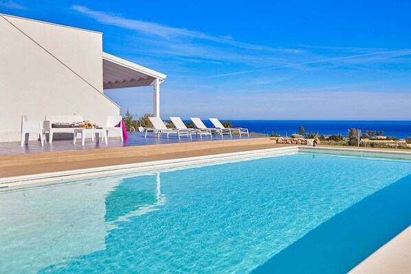 A poolside area with lounge chairs and a covered seating area, overlooking a clear blue sky and ocean.