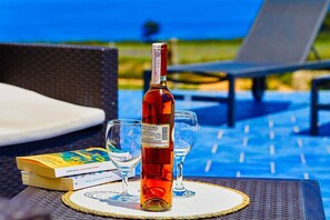 Une bouteille de vin, deux verres et un livre sur une table, près d’un fauteuil de salon installé au bord de la piscine.