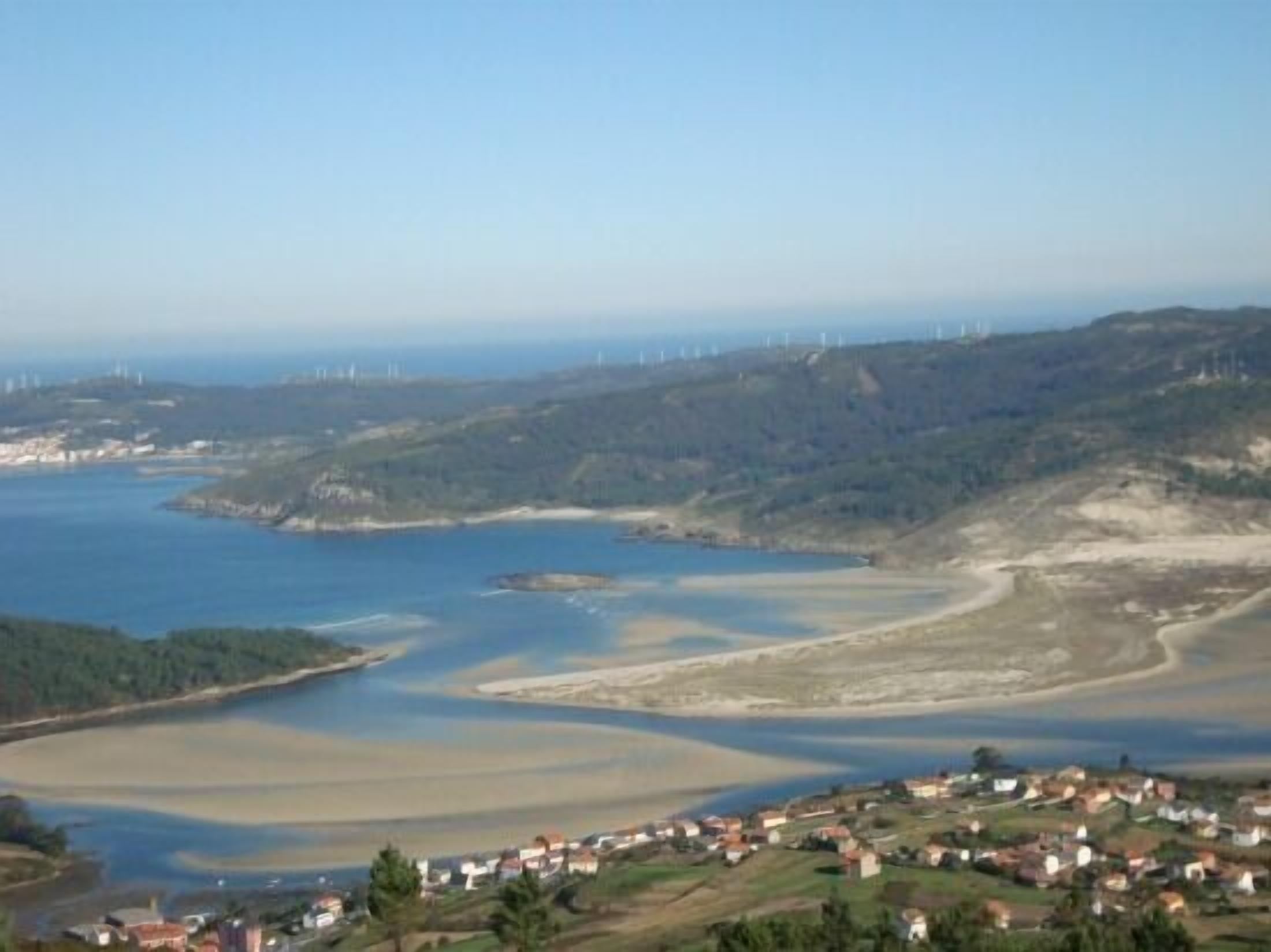 Un paisaje costero con un río, casas y aerogeneradores en la distancia.