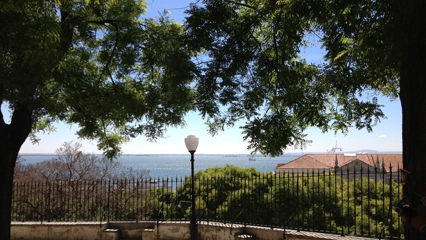A paved pathway with a lamp post, a metal fence, and a view of a body of water and buildings in the distance. - Riverview Bright Studio @Graça. New Deco 2023! (Lisboa)