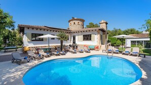 A swimming pool with lounge chairs and umbrellas, a stone tower, and a white building with shutters.