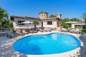 A swimming pool with lounge chairs and umbrellas, a stone tower, and a white building with shutters.