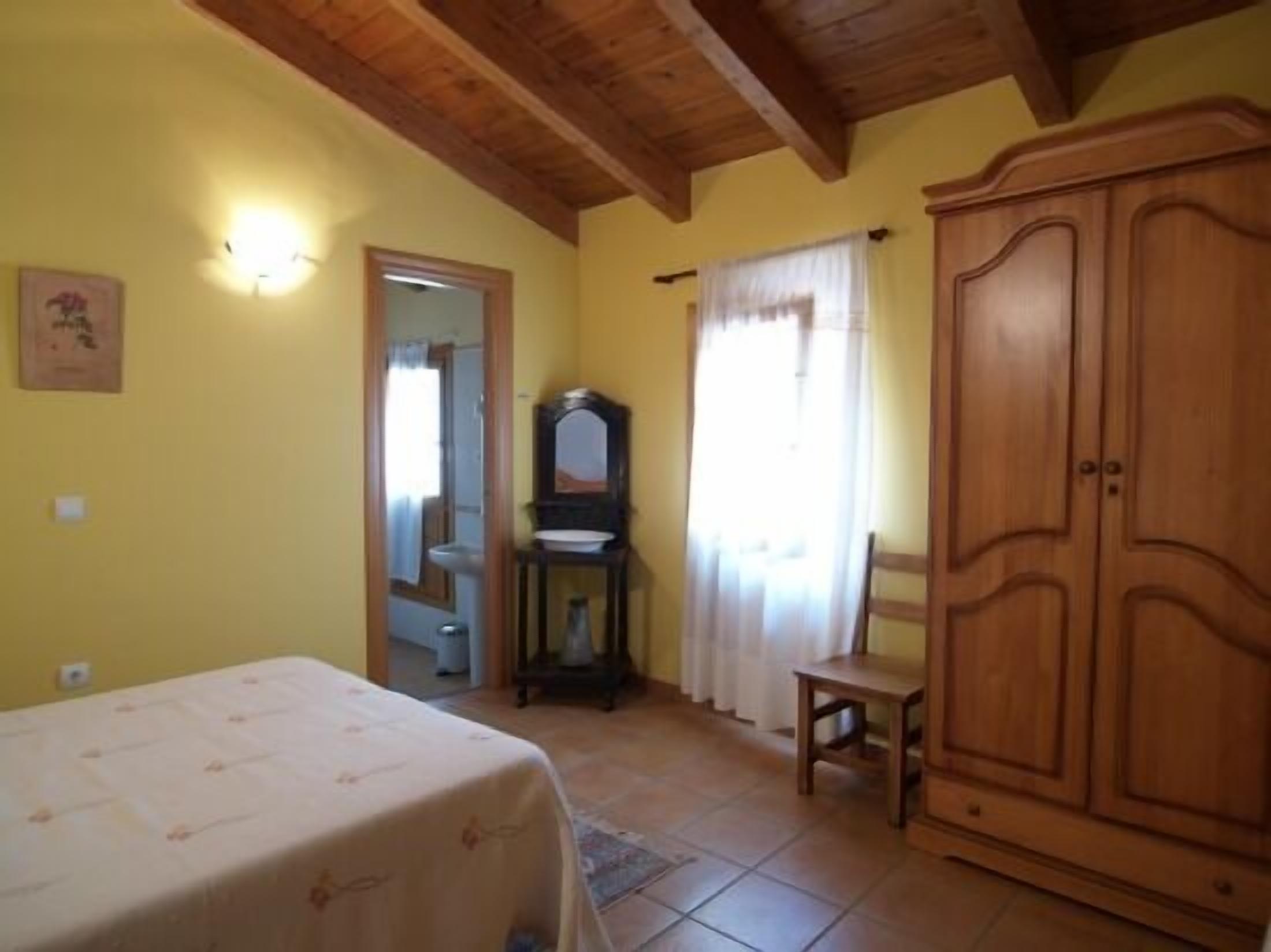 Une pièce comprenant un lit, une armoire en bois, une petite table, un miroir et une fenêtre avec des rideaux.