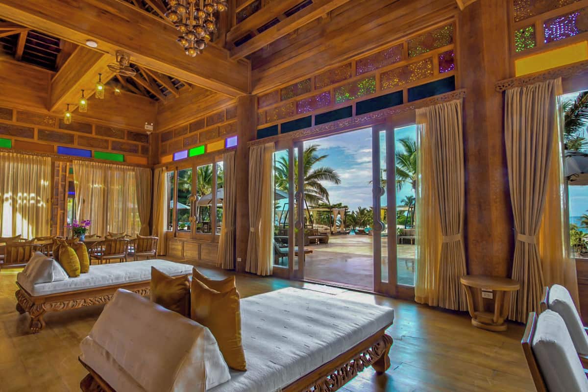 A spacious room with a large bed, a chair, and a table. There are large windows with curtains, and a view of palm trees outside.