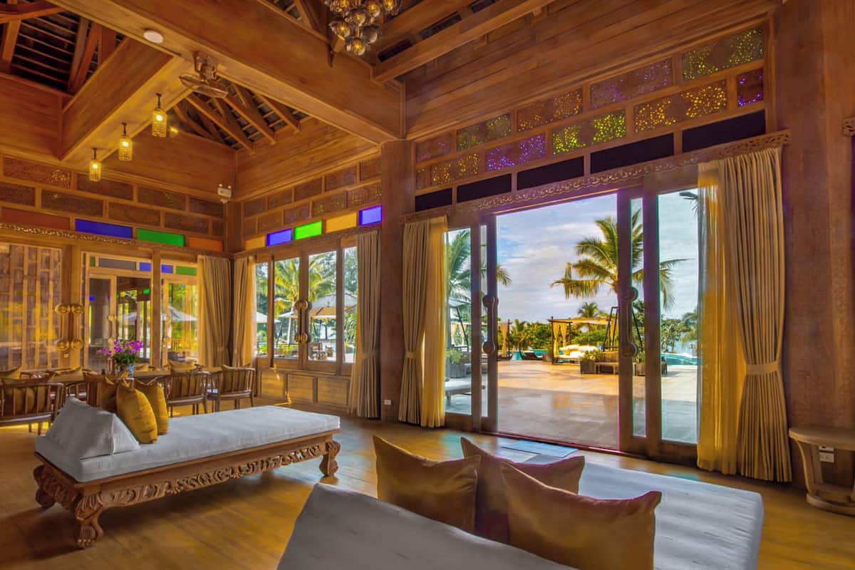 A spacious room with a large bed, a seating area with chairs, and a view of a pool and palm trees through large windows.