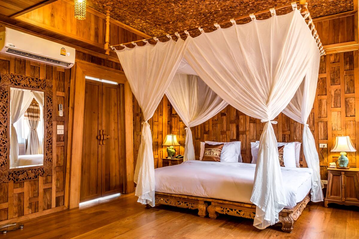 A four-poster canopy bed with a wooden headboard, a bedside table with a lamp, and a mirror with curtains.