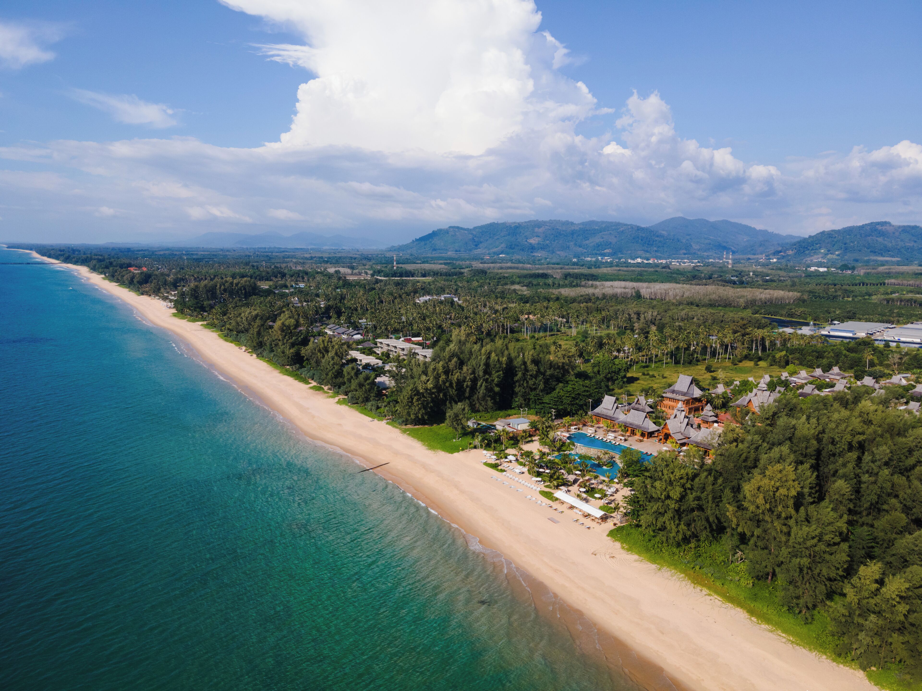 An aerial view of a coastal resort with a beach, a swimming pool, and surrounding forested areas.