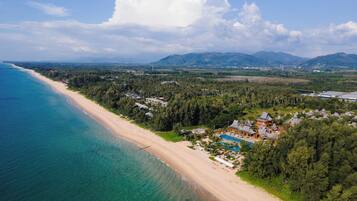 An aerial view of a coastal resort with a beach, a swimming pool, and surrounding forested areas.