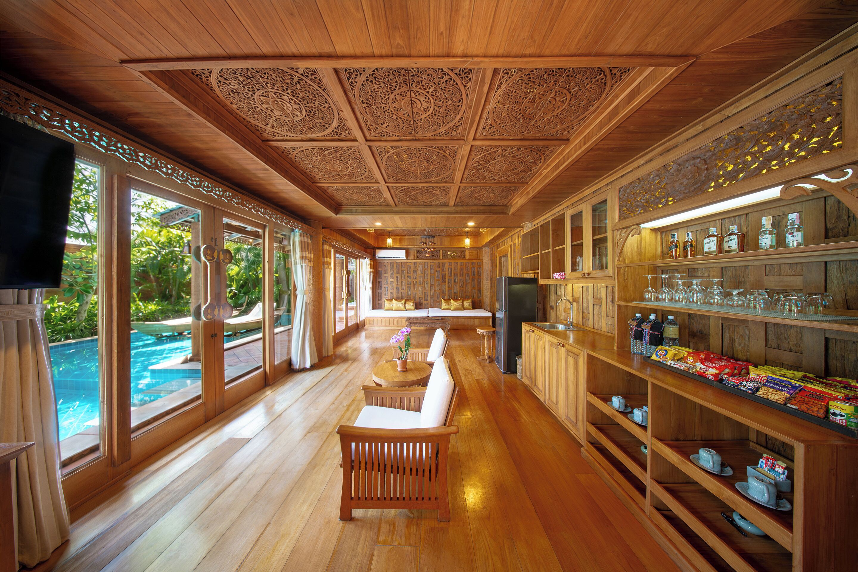 Une pièce spacieuse dotée d’un plancher en bois, d’un coin repas avec une table et des chaises, d’un bar avec des étagères remplies de bouteilles, et offrant une vue sur une piscine grâce à de grandes fenêtres.