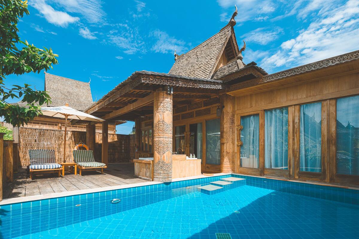 A wooden villa with a swimming pool, a covered patio with chairs, and large windows.