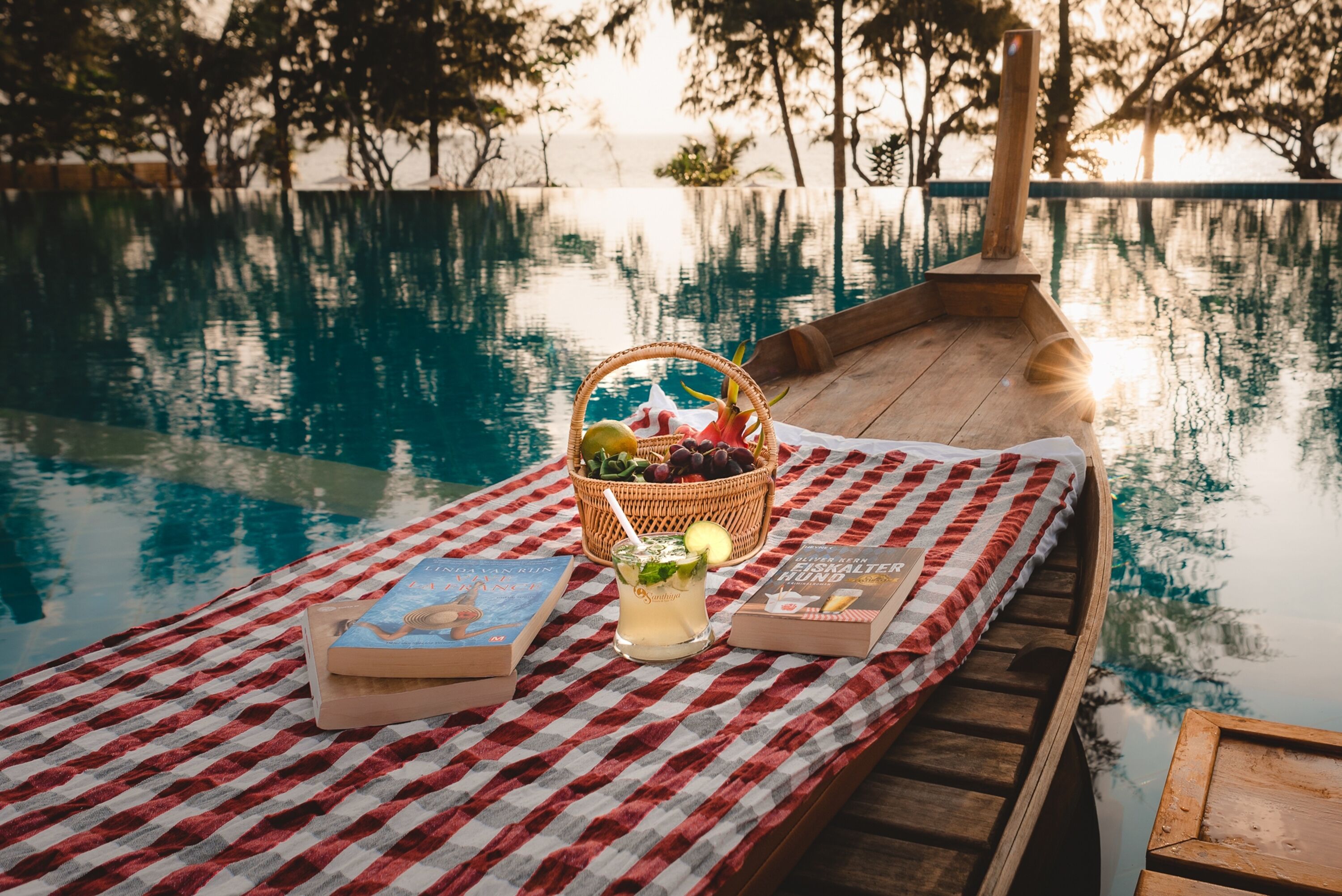 Un bateau en bois, une nappe à carreaux, un panier de fruits, une boisson et des livres, le tout placé au bord de la piscine.