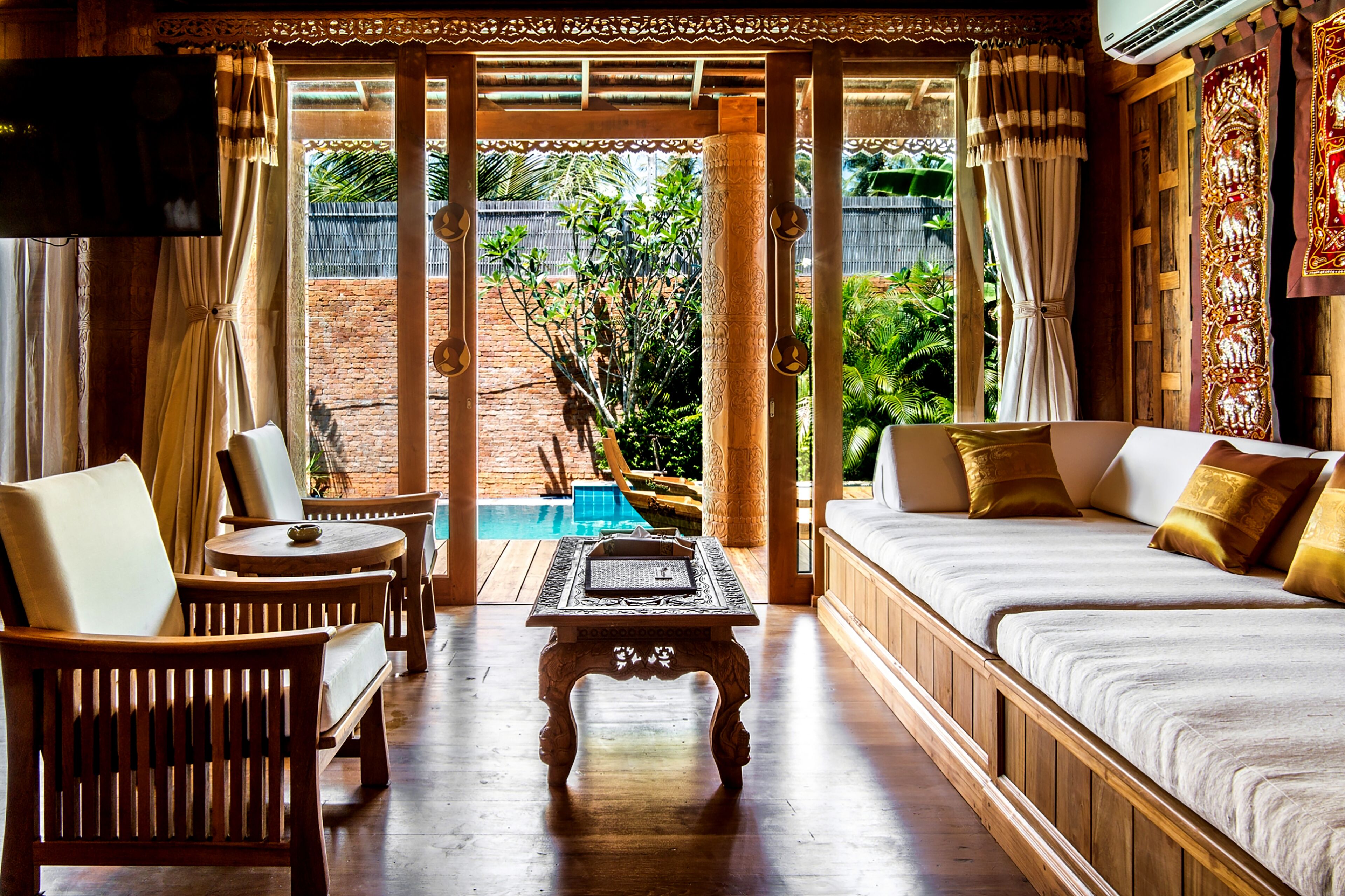 A room with a wooden sofa, a small table, and a TV. There is a view of a swimming pool and greenery outside.