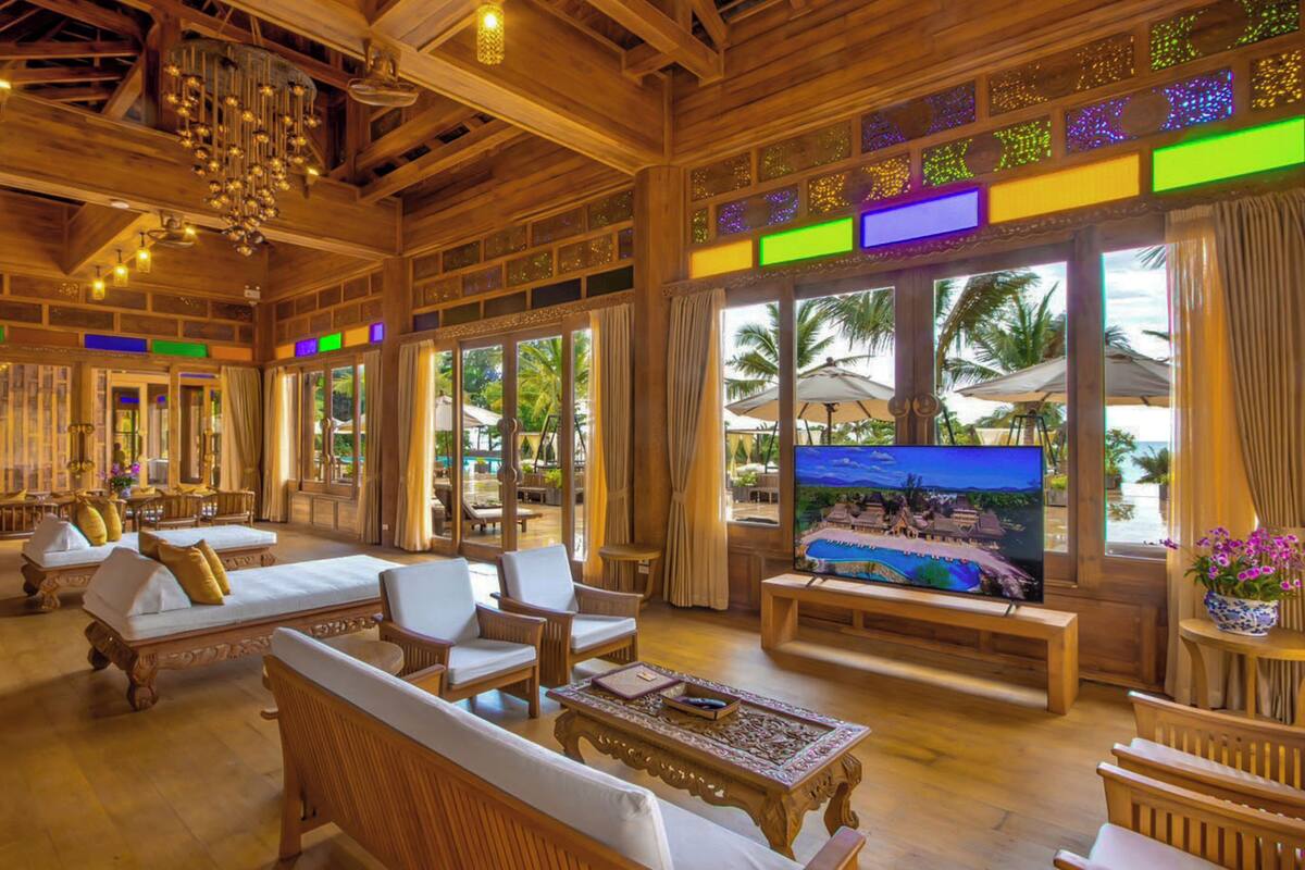 A spacious room with wooden furniture, a television, and large windows overlooking a beach.
