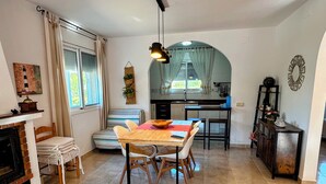A cozy living room with a fireplace, a dining area with a wooden table and chairs, a kitchen with a large window, and a decorative mirror on the wall.
