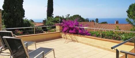 A balcony with a view of the sea, a pergola with purple flowers, and a tiled floor.