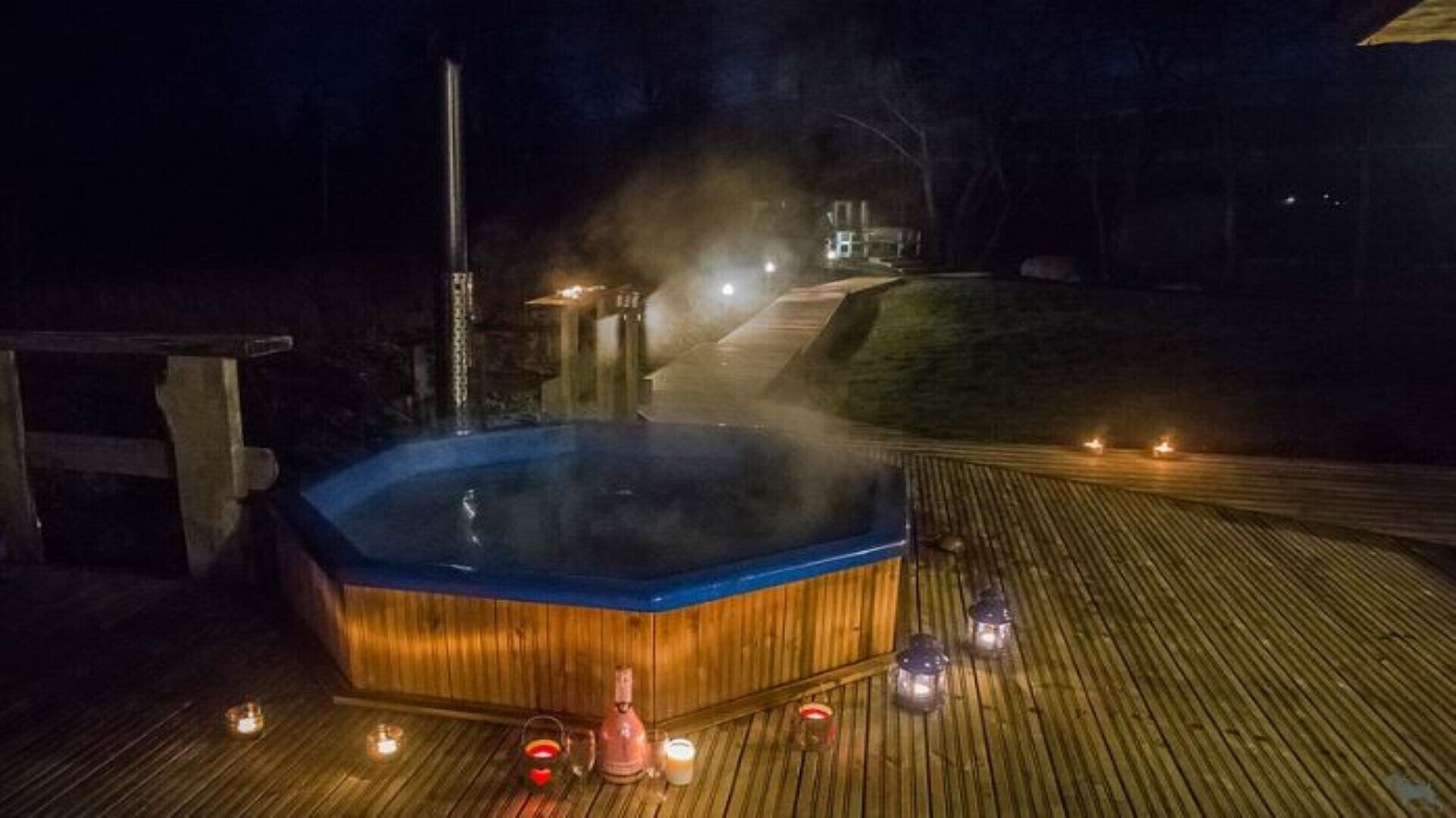 Uma banheira de hidromassagem numa varanda de madeira à noite, com ambiente enevoado.