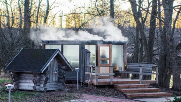 A cabin with a chimney emitting smoke, a wooden deck, and a log structure nearby. - Kivikoski riverside cottage in Pyhtää for 5-8 people (Pyhtää)