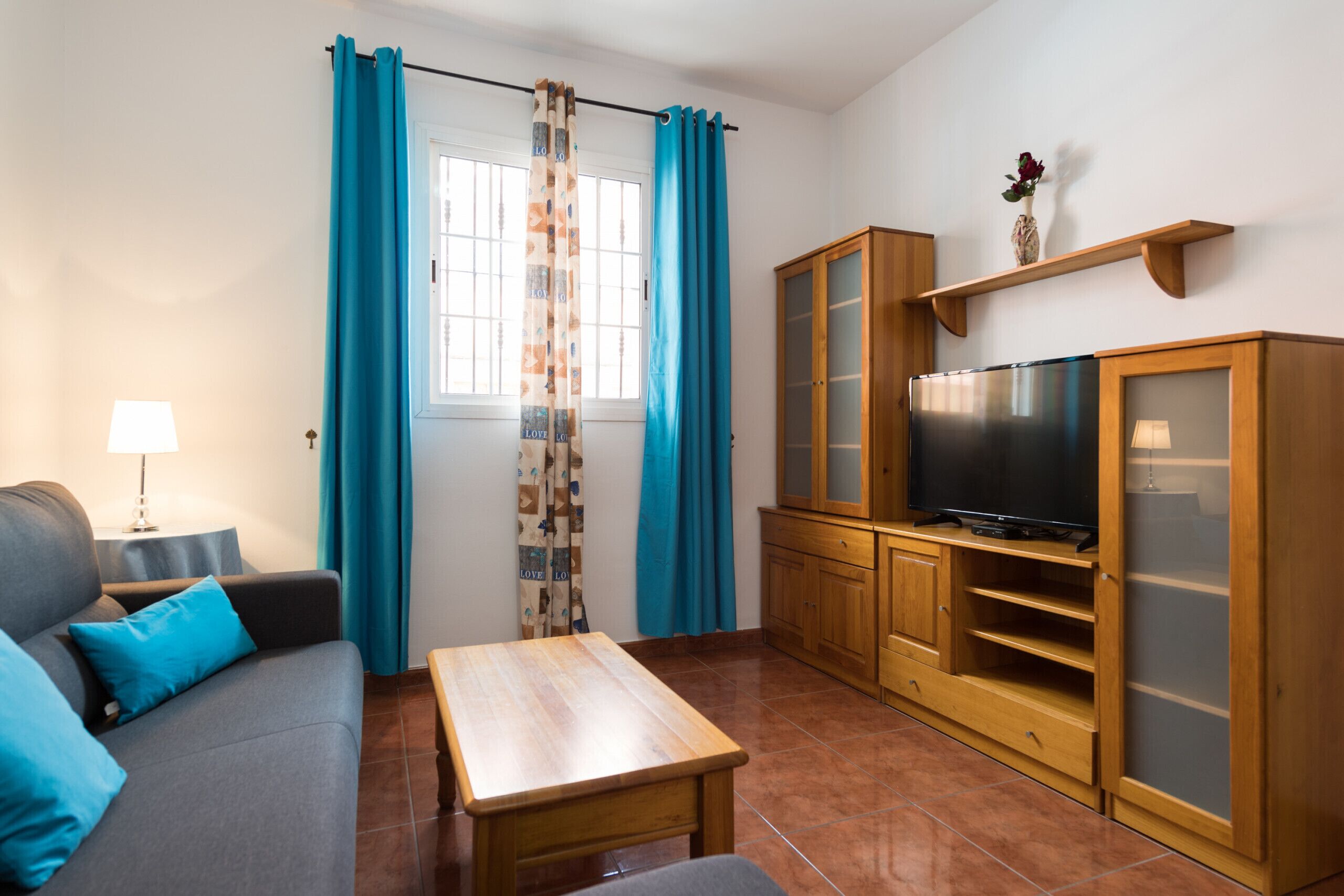 A living room with a sofa, a coffee table, a TV on a wooden cabinet, and a window with blue curtains.
