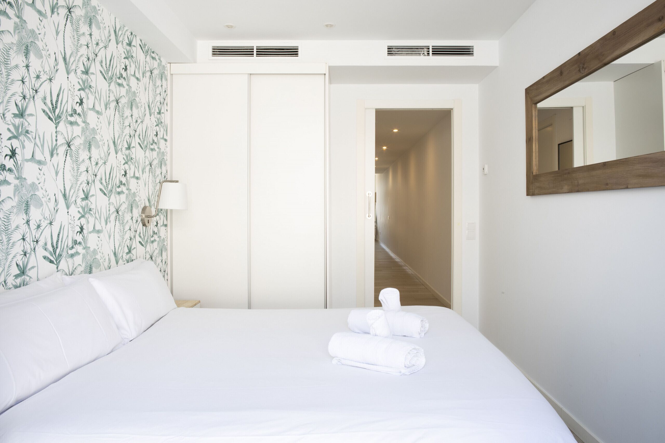 Ein Hotelzimmer mit einem Bett, weißen Leinentüchern, floralem Wandpapier, einem spiegelnden Holzrahmen und einem zur nächsten Tür führenden Flur.