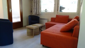 A room with a blue chair, a wooden table, an orange sofa, and a window with a view of the outside. - Detached chalet on the slopes and near the village, for winter sports & summer holidays (La Norma)