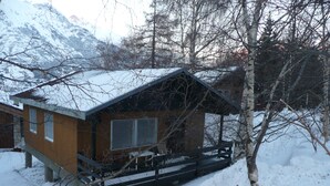 A wooden cabin with a porch, surrounded by snow and trees, with mountains in the background. - Detached chalet on the slopes and near the village, for winter sports & summer holidays (La Norma)