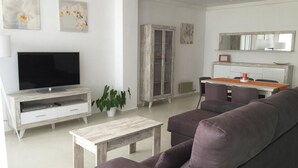 Een moderne woonkamer met een grijze bank, een salontafel, een televisie op een wit kastje, en een eethoek met een tafel en stoelen.