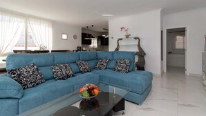 Un salon moderne avec un canapé d’angle bleu, des coussins aux motifs noir et blanc, une table basse en verre sur laquelle on aperçoit des fruits, et une cuisine en arrière-plan.
