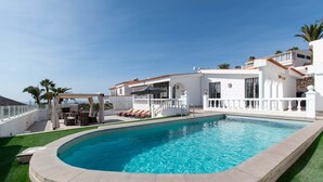 Une piscine entourée d’un bâtiment blanc aux bords incurvés, d’un patio couvert et de palmiers.
