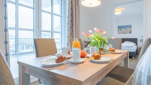 Une table de petit-déjeuner dressée avec des assiettes, des tasses et des viennoiseries, un vase rempli de fleurs et une vue sur l’extérieur grâce à de grandes fenêtres.