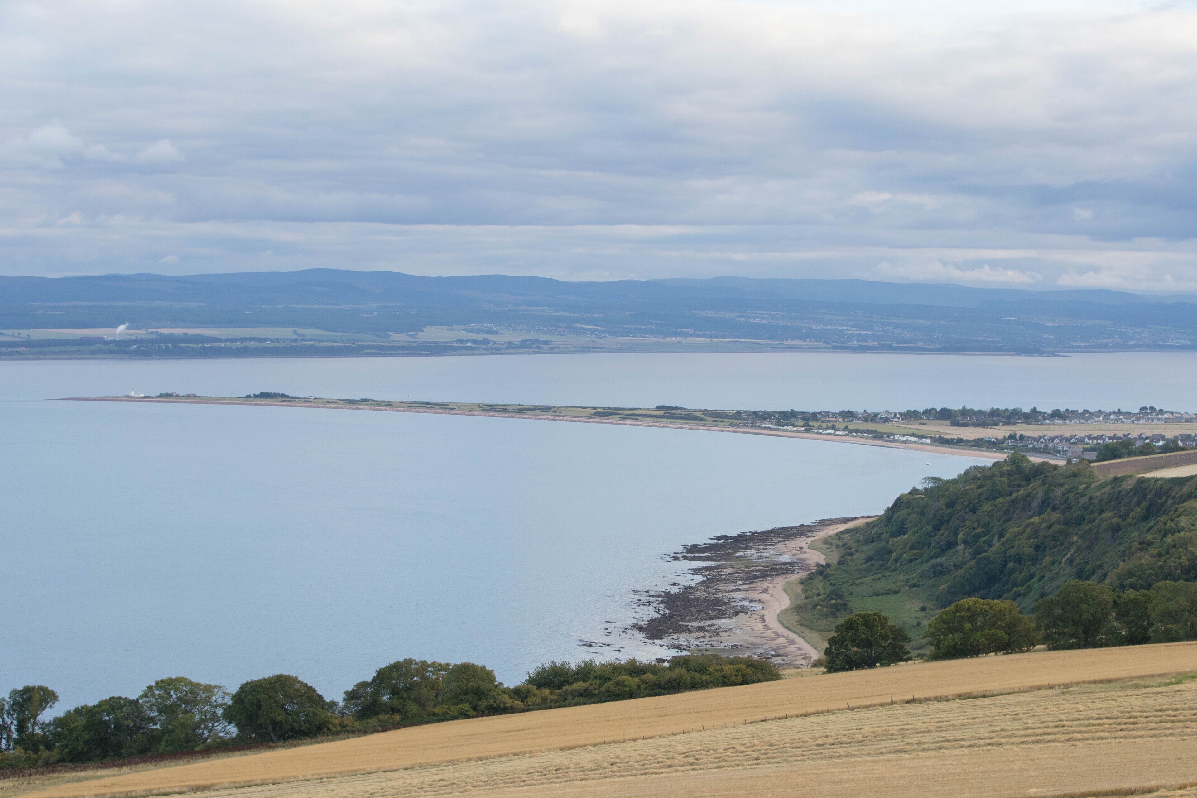 Un paysage côtier avec une plage, des champs et des collines à l’horizon.