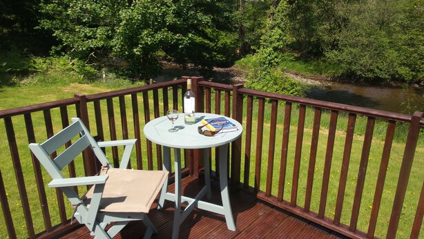 A deck with a table and a chair, overlooking a river and greenery. - Beautiful river front self catering holiday lodge. (Builth Wells)