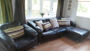A living room with a black leather sectional sofa, a wooden floor, and a window with curtains. - Beautiful river front self catering holiday lodge. (Builth Wells)