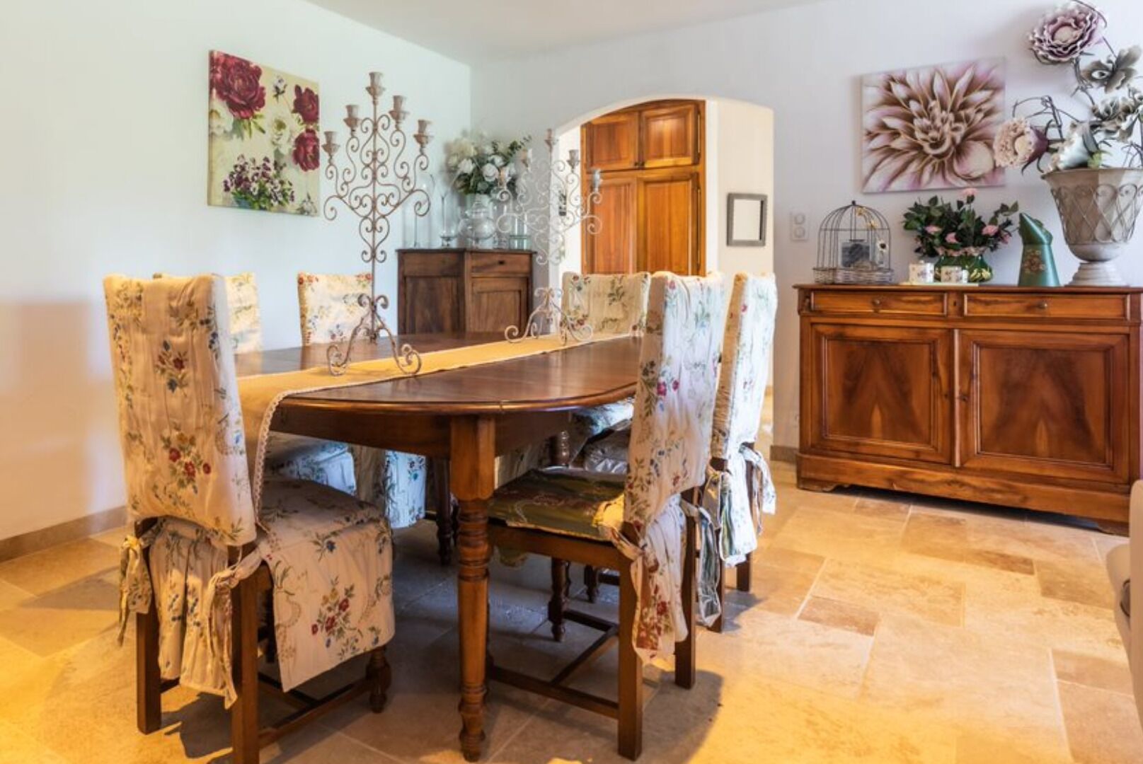 A dining area with a wooden table and floral-patterned chairs, a wooden cabinet, and floral artwork on the walls.